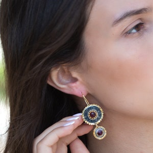 May include: Gold dangle earrings with a black and gold circular design and a red gemstone. The earrings have a smaller red gemstone dangling below the larger circular design.