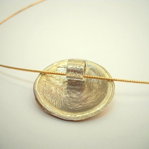 Puede incluir: Un colgante de plata con una superficie texturizada cuelga de una fina cadena de oro. El colgante tiene un peque&ntilde;o anillo de plata en la parte posterior con la inscripci&oacute;n "HW".