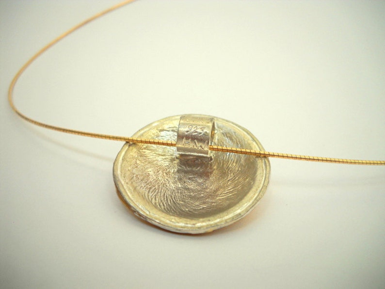 Puede incluir: Un colgante de plata con una superficie texturizada cuelga de una fina cadena de oro. El colgante tiene un peque&ntilde;o anillo de plata en la parte posterior con la inscripci&oacute;n "HW".