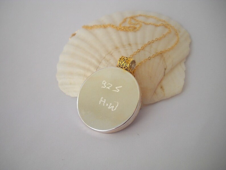 May include: A silver round pendant necklace with a gold chain. The pendant has the inscription "925" and "H-W" on the front.