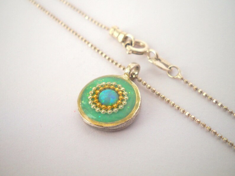 May include: A silver chain necklace with a round pendant featuring a green background, a gold rim, and a blue opal surrounded by gold and green beads.