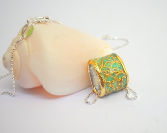 May include: A silver necklace with a pendant. The pendant is a cylinder shape with a gold-colored rim and a light green center with a gold filigree design. The necklace is on a white background.