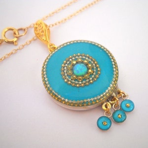 Puede incluir: Un collar de cadena de oro con un colgante redondo que presenta un centro azul turquesa con una piedra de &oacute;palo blanco y detalles dorados. El colgante tiene tres peque&ntilde;os abalorios azules turquesa que cuelgan de la parte inferior.