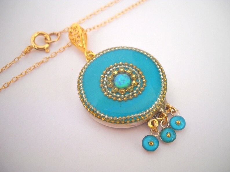 Puede incluir: Un collar de cadena de oro con un colgante redondo que presenta un centro azul turquesa con una piedra de &oacute;palo blanco y detalles dorados. El colgante tiene tres peque&ntilde;os abalorios azules turquesa que cuelgan de la parte inferior.