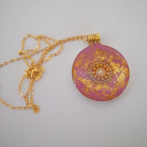 May include: A pink and gold round pendant necklace with a pearl in the center. The pendant has a gold leaf design and is attached to a gold chain.