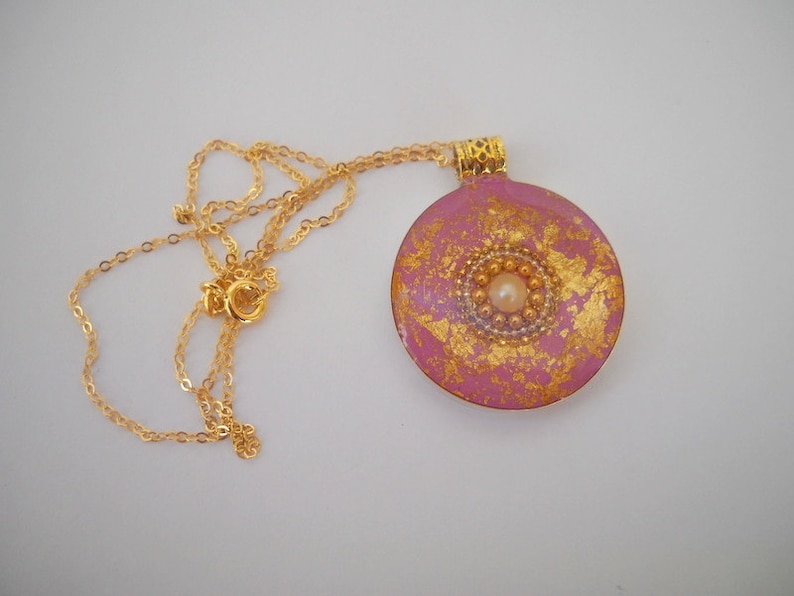 May include: A pink and gold round pendant necklace with a pearl in the center. The pendant has a gold leaf design and is attached to a gold chain.