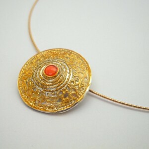 Puede incluir: Un collar con un colgante redondo de oro y plata, con un dise&ntilde;o de filigrana y una peque&ntilde;a piedra roja en el centro.