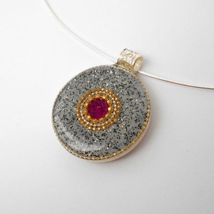 May include: A silver chain necklace with a round pendant. The pendant is made of gray glitter resin with a gold rim and a pink gem in the center.