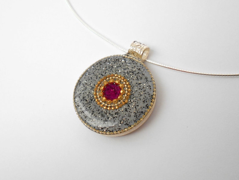 May include: A silver chain necklace with a round pendant. The pendant is made of gray glitter resin with a gold rim and a pink gem in the center.