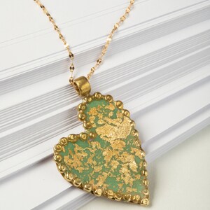 Puede incluir: Un collar de cadena dorada con un colgante en forma de corazón verde. El colgante está decorado con pan de oro y tiene un borde de cuentas.