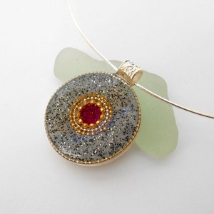 May include: A silver chain necklace with a round pendant featuring a gray and black glitter background with a gold and red center. The pendant is attached to the chain with a silver clasp.