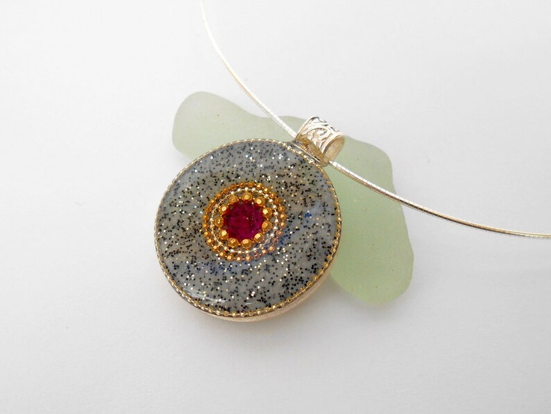 May include: A silver chain necklace with a round pendant featuring a gray and black glitter background with a gold and red center. The pendant is attached to the chain with a silver clasp.