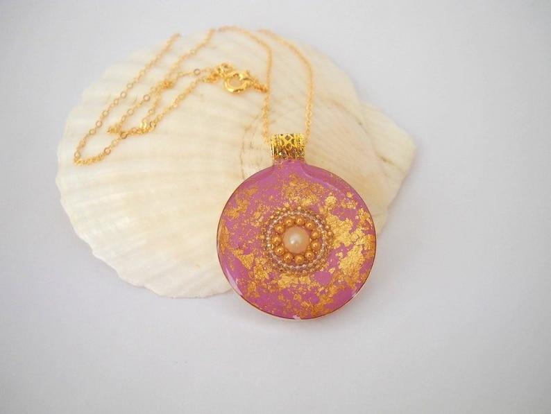 May include: A pink and gold round pendant necklace with a pearl and gold bead design. The necklace is on a gold chain.