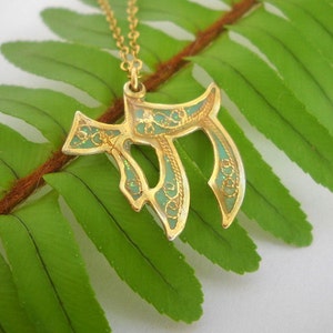 May include: A gold pendant necklace with a green enamel design in the shape of the Hebrew letter Chai. The pendant is on a fern leaf background.