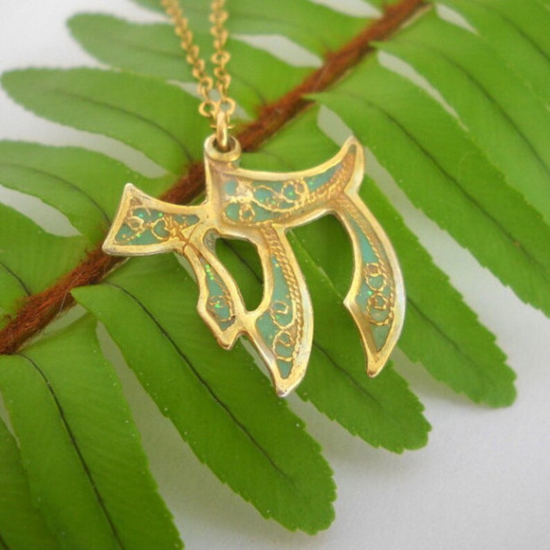 May include: A gold pendant necklace with a green enamel design in the shape of the Hebrew letter Chai. The pendant is on a fern leaf background.
