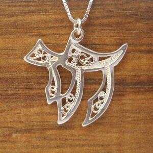May include: A silver pendant necklace featuring a delicate filigree design in the shape of the Hebrew letter Chai. The pendant is suspended from a silver chain.