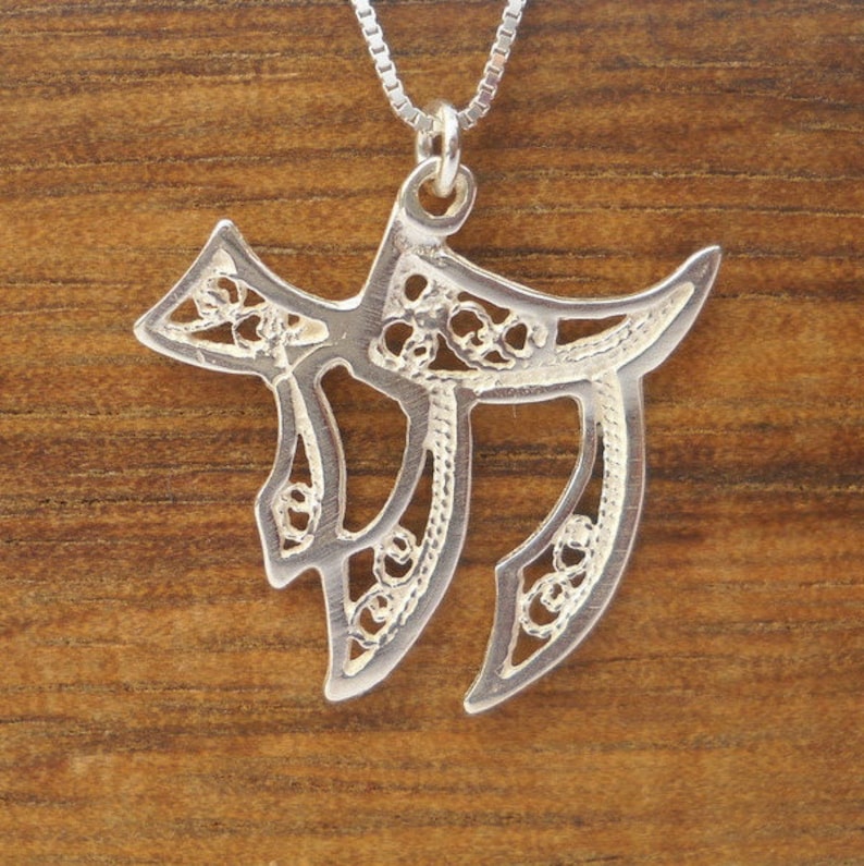 May include: A silver pendant necklace featuring a delicate filigree design in the shape of the Hebrew letter Chai. The pendant is suspended from a silver chain.