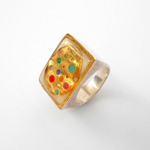 May include: A silver ring with a square-shaped top. The top is made of white enamel and features a gold-colored design of a circle with colorful dots.