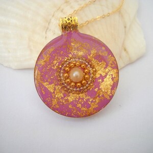 May include: A pink round pendant necklace with gold flecks and a pearl in the center. The pendant is on a gold chain.