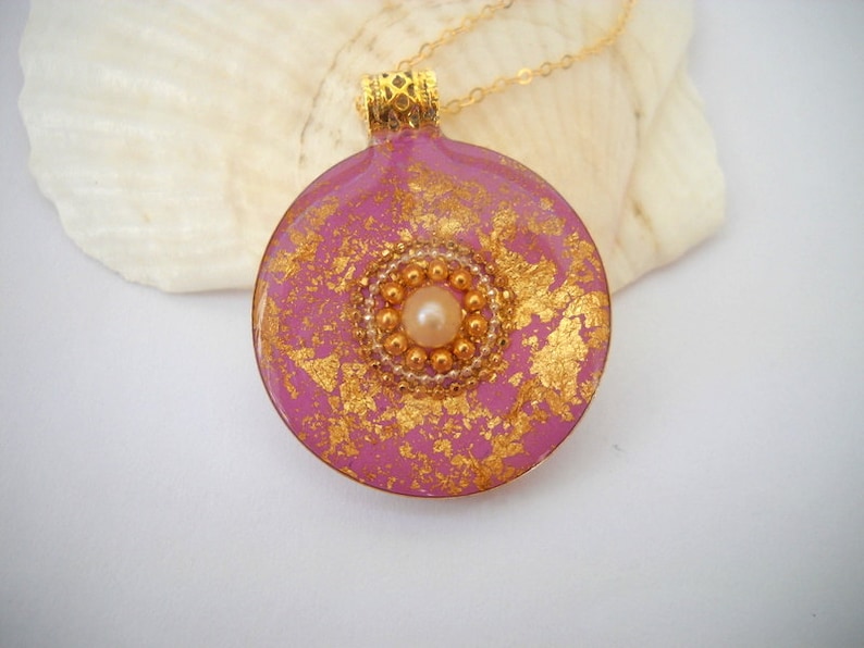 May include: A pink round pendant necklace with gold flecks and a pearl in the center. The pendant is on a gold chain.
