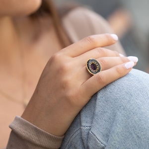 May include: Oval-shaped ring with a dark background and a central purple stone. The ring has a gold-colored band and is adorned with small, gold-colored beads around the purple stone. The ring is on a person's finger.