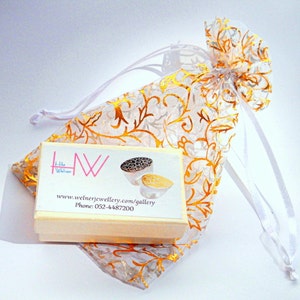 May include: A small white box with gold lettering and a gold and black ring inside. The box is sitting on a white and gold patterned fabric bag. The text on the box reads "I Like Welner Jewellery.com/gallery Phone: 052-4487200"