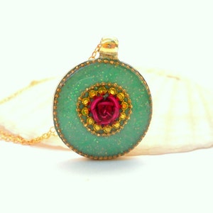 Puede incluir: Un collar con un colgante redondo verde y dorado, adornado con una rosa roja en el centro. El colgante es brillante.
