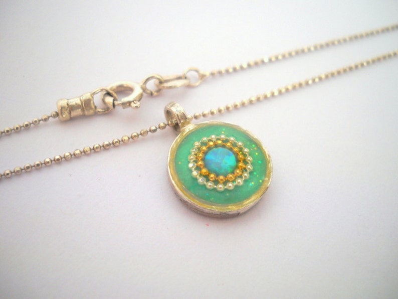 May include: A silver chain necklace with a round pendant featuring a green background, gold and white beads, and a blue opal in the center.