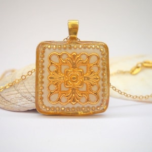 Puede incluir: Un collar de oro con un colgante cuadrado blanco con un diseño de filigrana dorada. El colgante está en una cadena de oro.