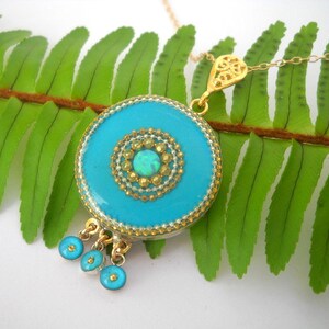 Puede incluir: Un collar con un colgante azul turquesa con un dise&ntilde;o de filigrana dorada y una peque&ntilde;a piedra de &oacute;palo en el centro. El colgante est&aacute; rodeado por un borde de cuentas doradas y tiene tres peque&ntilde;as cuentas azules turquesa que cuelgan de la parte inferior.