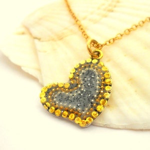 Puede incluir: Un collar de cadena dorada con un colgante en forma de corazón. El colgante está hecho de metal gris con un acabado brillante y un borde de pequeñas cuentas doradas.