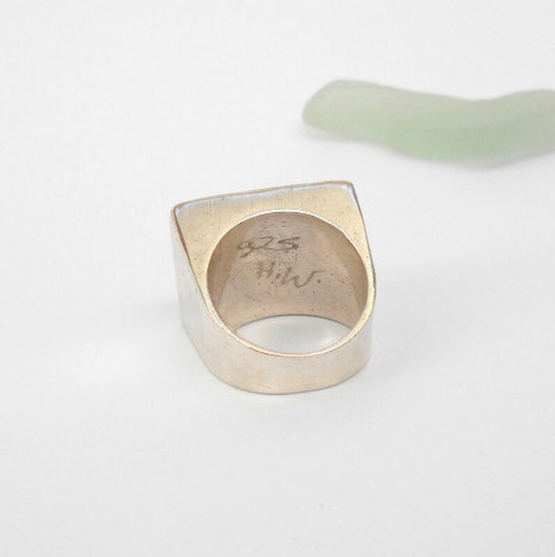 May include: A silver ring with a square top and a rounded band. The inside of the band is stamped with "925 Hilv".