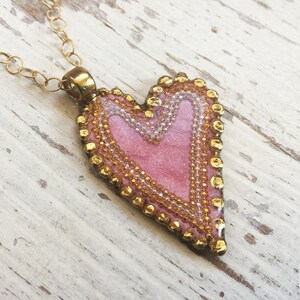 Puede incluir: Un collar con un colgante en forma de corazón rosa, una cadena dorada y un borde de cuentas. El colgante tiene un acabado brillante y texturizado.