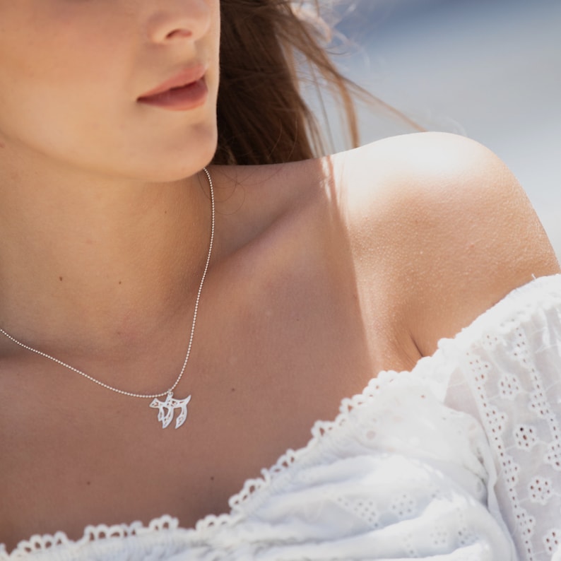 May include: A silver necklace with a delicate pendant featuring a stylized tree of life design. The necklace is worn by a person with a white, off-the-shoulder top.