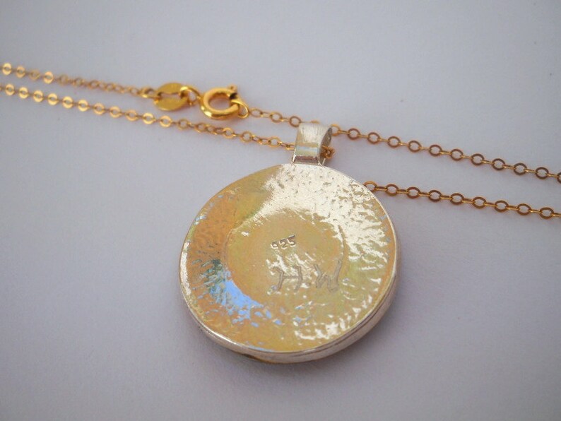 May include: A silver pendant necklace with a round, hammered silver pendant. The pendant has a small inscription on the back that reads "925". The necklace is attached to a gold chain.