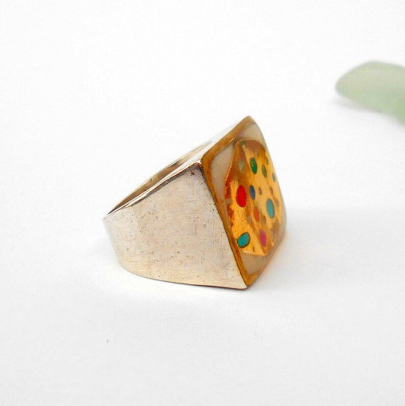 May include: A silver ring with a square-shaped top. The top is white with a gold design of colorful dots.