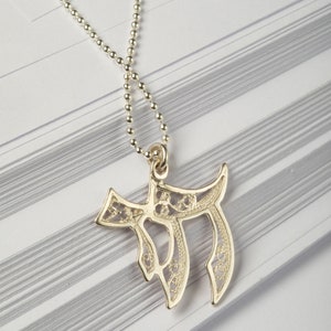 May include: A silver chain necklace with a delicate, filigree pendant in the shape of the Hebrew letter Chai. The pendant is intricately designed with a floral pattern.