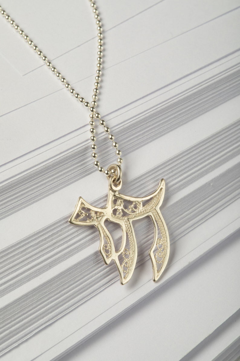 May include: A silver chain necklace with a delicate, filigree pendant in the shape of the Hebrew letter Chai. The pendant is intricately designed with a floral pattern.