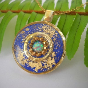 May include: A gold pendant necklace with a blue and gold round pendant. The pendant has a gold circle with a small opal gemstone in the center. The pendant is surrounded by gold flakes.
