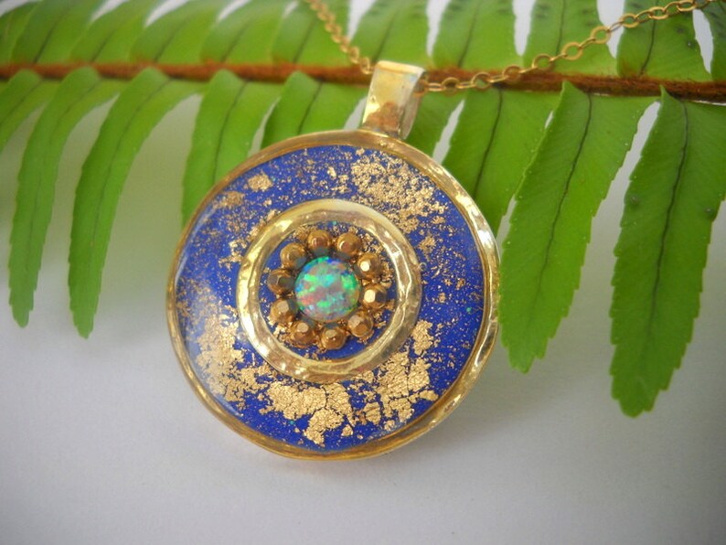 May include: A gold pendant necklace with a blue and gold round pendant. The pendant has a gold circle with a small opal gemstone in the center. The pendant is surrounded by gold flakes.