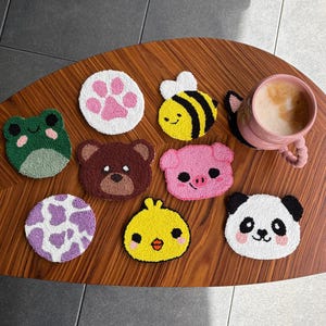 Könnte beinhalten: Eine Sammlung handgefertigter Untersetzer mit Tiermotiven, darunter ein Frosch, ein Bär, eine Biene, ein Schwein, ein Panda und ein Küken. Die Untersetzer liegen auf einem Holztisch, neben einer rosa Kaffeetasse.