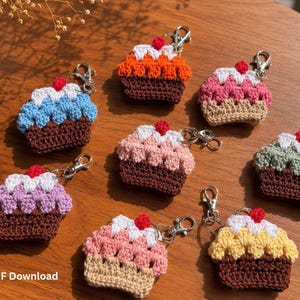 Puede incluir: Una colección de siete llaveros de cupcakes tejidos a ganchillo, cada uno con una base marrón o beige, una cobertura texturizada de colores en azul, naranja, rosa, verde, morado o amarillo, una capa blanca y una pequeña cereza roja.