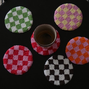 Könnte beinhalten: Draufsicht auf einen schwarzen Tisch mit sechs bunten, runden Untersetzern. Die Untersetzer haben ein Schachbrettmuster in Grün, Rosa, Orange, Schwarz und Gelb. In der Mitte steht eine braune Tasse.