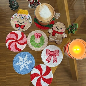 Könnte beinhalten: Eine Sammlung handgefertigter Weihnachtsuntersetzer mit Lebkuchenmann-, Schneeflocken- und Pfefferminz-Wirbel-Designs. Eine Kaffeetasse steht auf einem rot-grünen Untersetzer. Dekorative Weihnachtsbaum- und Weihnachtsmannfiguren sind ebenfalls vorhanden.