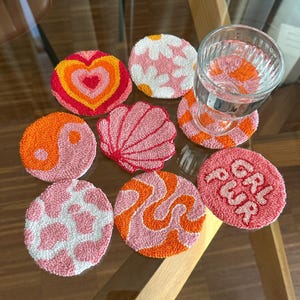 Könnte beinhalten: Eine Sammlung bunter, runder Untersetzer mit verschiedenen Designs, darunter ein Herz, eine Blume, ein Yin-Yang-Symbol, eine Muschel und ein Wirbelmuster. Ein Untersetzer hat den Text "GRL PWR". Die Untersetzer sind aus einem strukturierten Material.