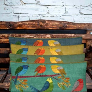 Peut inclure: Une pile de quatre coussins rectangulaires ornés d'oiseaux brodés perchés sur des branches. Les coussins présentent un dégradé de couleurs allant du bleu foncé au vert clair, avec des oiseaux bleus, orange et jaunes sur chaque pièce.