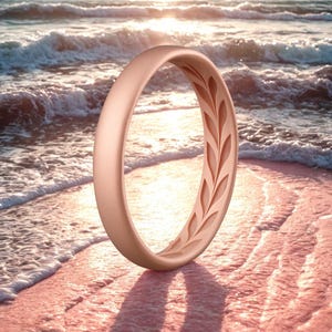 May include: A rose gold ring with a leaf pattern carved into the inner band. The ring is standing on a sandy beach with waves in the background, illuminated by the sun.
