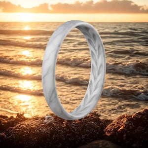 May include: A white marble-effect ring with a leaf pattern. The ring is set against a sunset over the sea. The ring has a minimalist design.