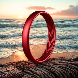 May include: A red ring with a leaf pattern on the inside, set against a beach scene with the ocean and a sunset. The ring is made of a solid material and is resting on a piece of driftwood.