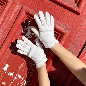Può includere: Un paio di guanti bianchi con un delicato bordo in pizzo smerlato. I guanti sono realizzati in materiale liscio. Lo sfondo è una porta rossa usurata.